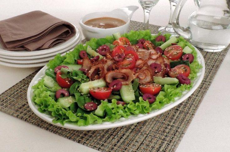 A Salada com rosbife está sendo servida em uma travessa branca. Os ingredientes estão dispostos no centro da salada, sendo eles o pepino, o rosbife, o tomate, a azeitona, o agrião e a rúcula.