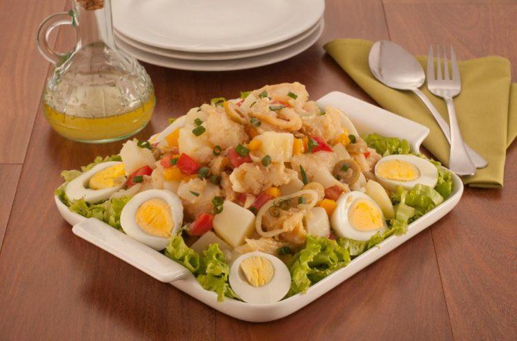 Foto da receita da salada de bacalhau e ovos, disposta em uma travessa de vidro branca. Ao fundo acompanham pratos, uma jarra de azeite, e 2 talheres em cima de de um guardanapo verde.