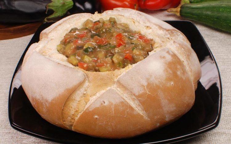A Sopa de berinjela está sendo servida dentro de um pão italiano. A Sopa possui pedaços visíveis de berinjela e tomate.