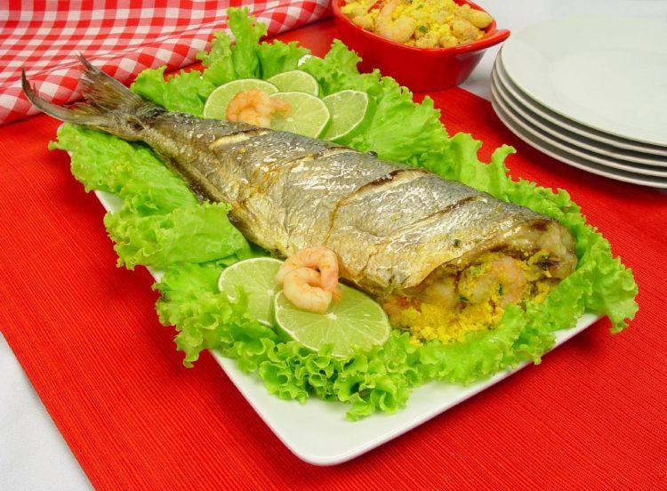 Foto com a receita de peixe assado recheado disposta em uma travessa branca, forrada com alface, camarão e limão. Ao fundo estão pratos brancos e uma tigela de camarão com farofa.