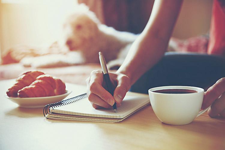 mulher escrevendo num caderno tomando cafe com cachorro ao fundo