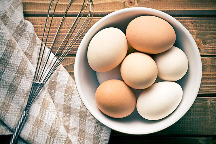 Ovos frescos de galinha na tigela com pano de prato e batedor de arame ao lado.