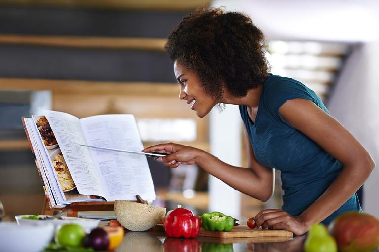 Mulher observando uma receita em um livro, com os ingredientes à mesa para prepará-la