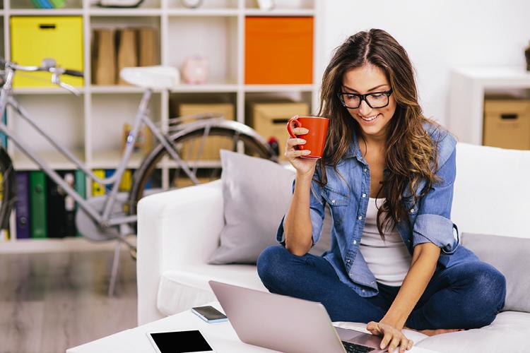 Mulher sentada no sofá com uma xícara de café na mão e sorrindo para o computador