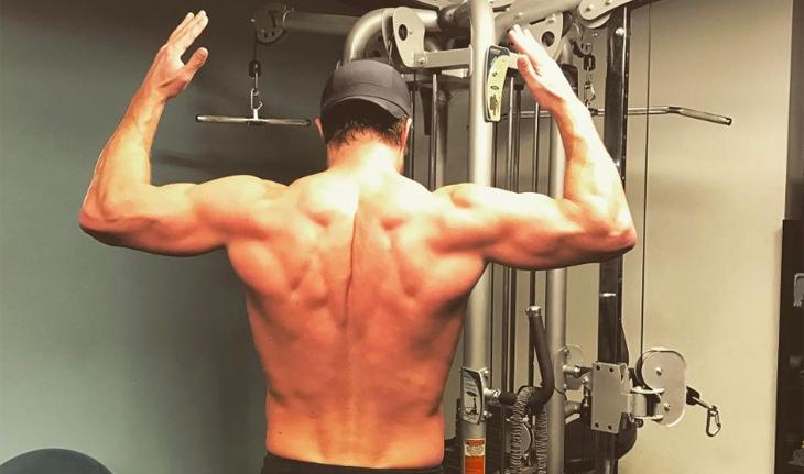 Stephen Amell, de costas e com os braços levantados, mostra os músculos na academia