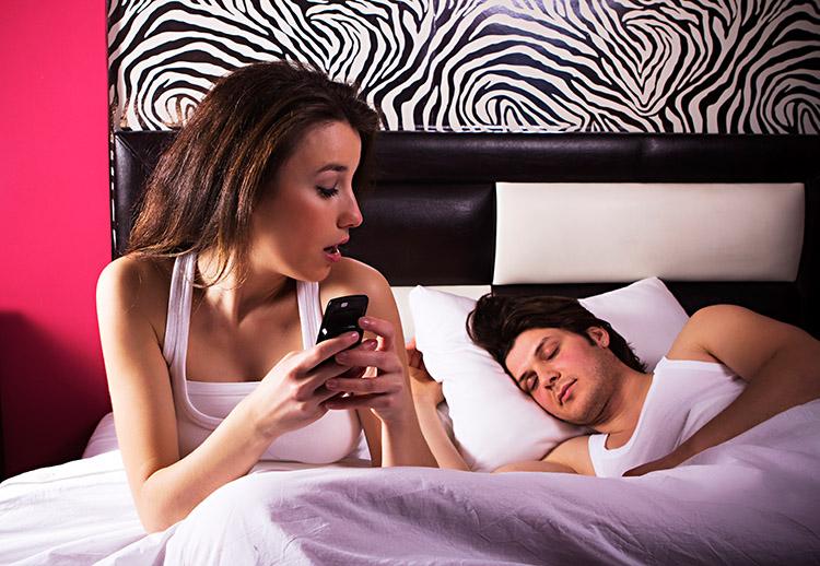 mulher sentada na cama mexendo no celular enquanto o homem está dormindo ao lado dela