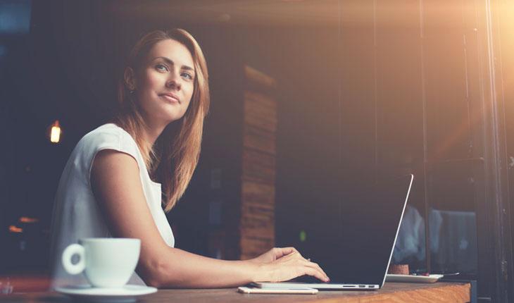 mulher mexendo no computador com uma xícara branca ao seu lado, olhando para frente e sorrindo