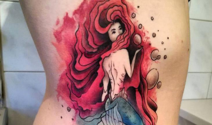Imagem de tatuagem na costela em aquarela de uma sereia de costas, olhando para trás, com o cabelo vermelho, longo, volumoso e ondulado, com cauda verde e bolhas ao seu redor