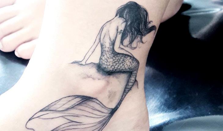 Imagem de tatuagem em preto e branco de uma sereia sentada em uma pedra fora da água olhando para baixo