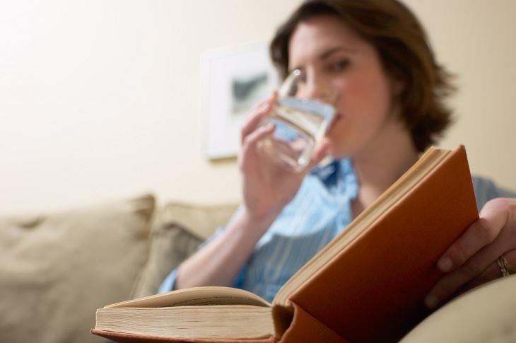 mulher bebendo água enquanto lê um livro no sofá