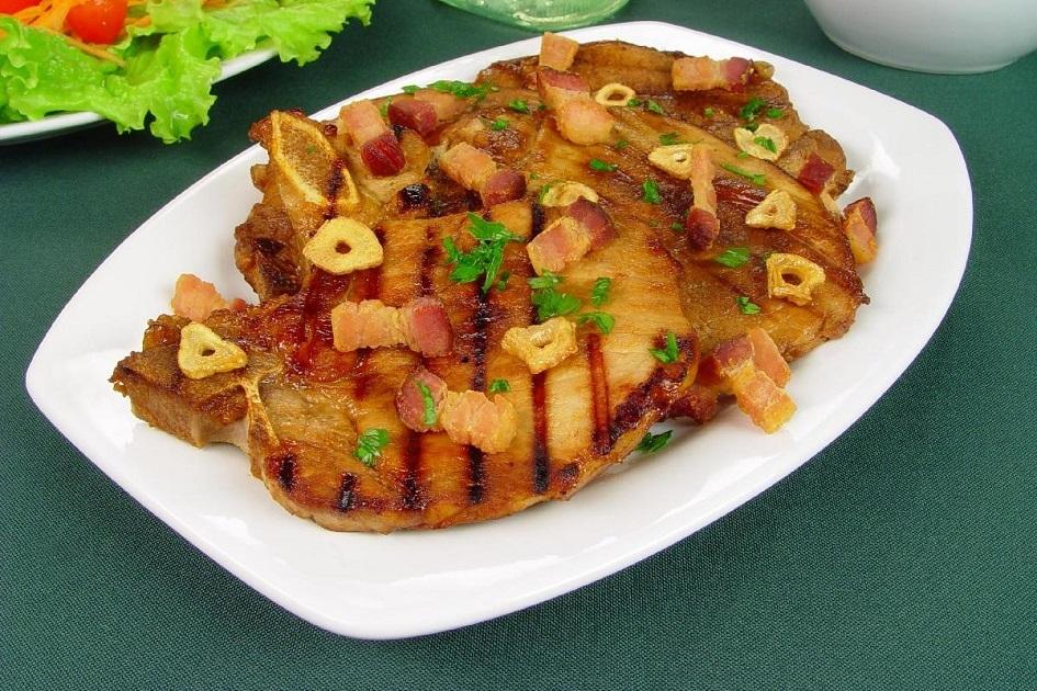 A bisteca de porco ao alho e bacon está sobre um prato branco oval e os pedacinhos do bacon e do alho estão por cima