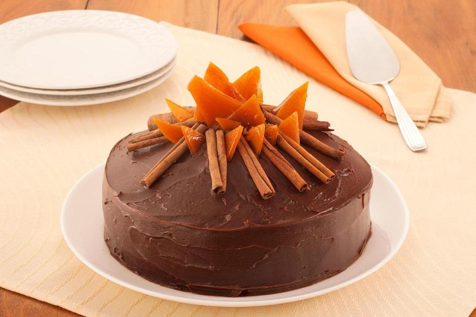 Na foto, o bolo fogueira está em um prato de servir grande. O bolo tem cobertura de chocolate e decoração de canela em pau e caramelos quebrados, simulando uma fogueira de festa junina.