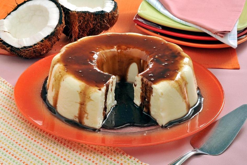Na foto, o manjar prestígio está em um prato redondo grande e tem um pedaço cortado. O manjar tem calda de chocolate despejada em toda a volta.