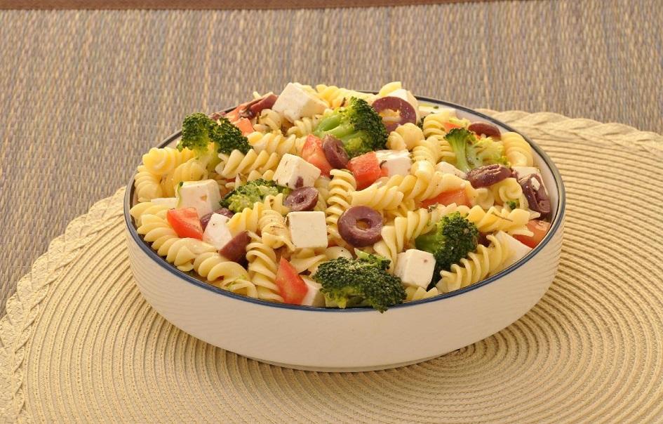 A salada de macarrão e brócolis está dentro de uma travessa redonda e branca e é possível ver todos os ingredientes, como azeitona, queijo, tomate e o brócolis