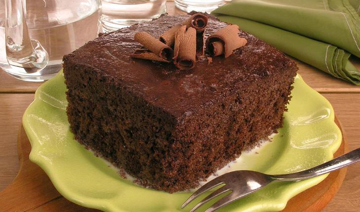 bolo chocolate molhado servido em um pratinho verde com um garfo de sobremesa, com copos e uma jarra de vidro com água ao fundo