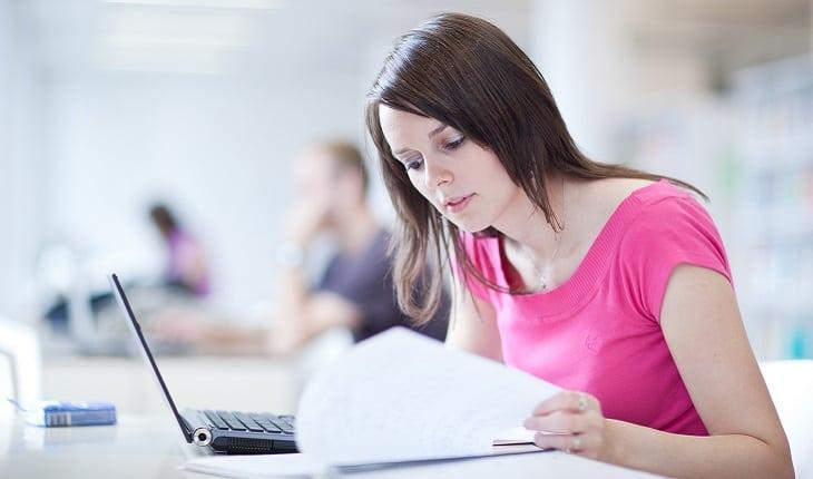 A foto mostra uma menina com camiseta rosa estudando com um notebook e lendo suas anotações