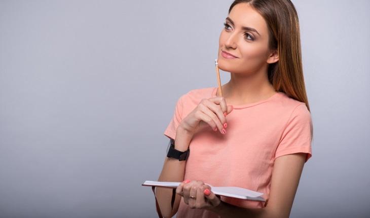 A foto mostra uma mulher com um caderno na mão, olhando para cima com um lápis encostado no queixo tentando encontrar motivações, um fator importante para descobrir como controlar as emoções