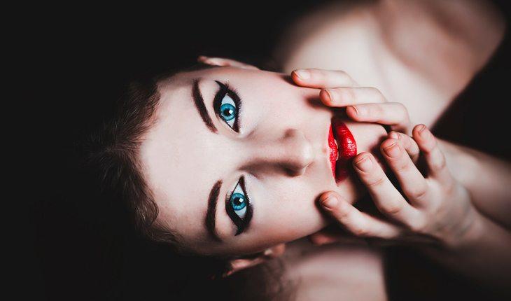 mulher de olhos azuis olhando para cima com a mão no rosto maquiado com batom vermelho e olho delineado gatinho