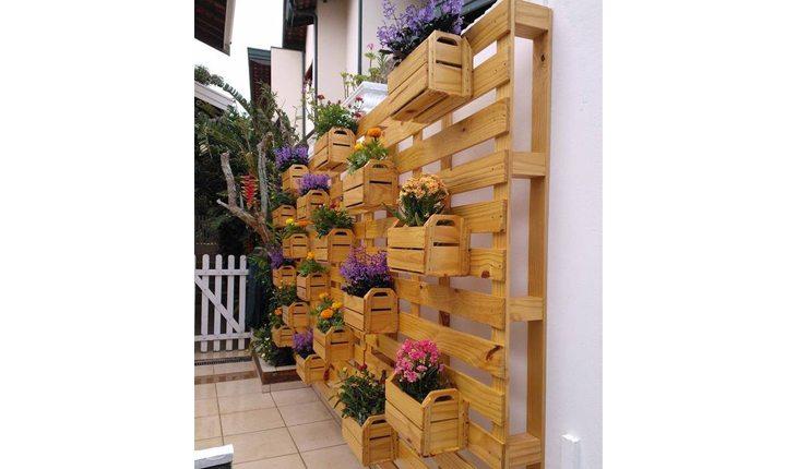 Na foto há um jardim vertical que ocupa toda uma parede feita com paletes. Em alguns pontos das paletes há vasinhos compridos de madeira onde estão plantadas plantas com flores.