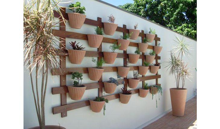 Na foto há um suporte para jardim na parede feito com ripas de madeira na horizontal. Os vasos são de barro e há plantas.