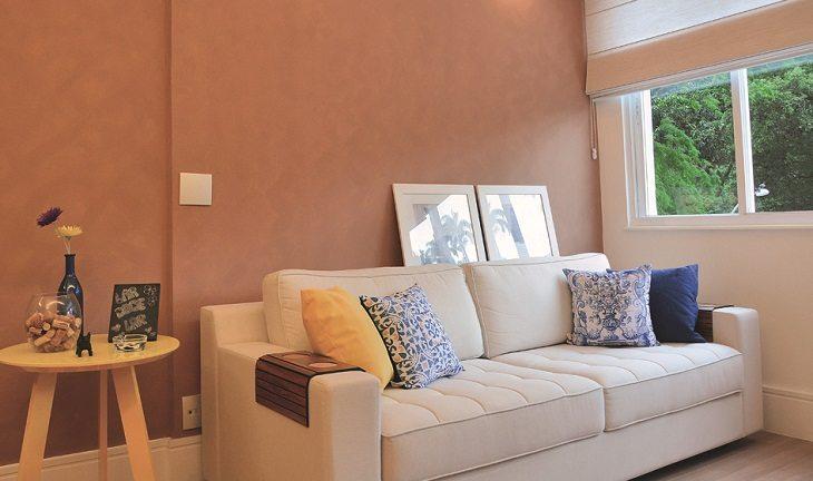 Sala com visual repaginado com tons neutros, parede em rosé, sofá branco com algumas almofadas coloridas, no canto esquerdo uma mesa rústica de madeira com alguns objetos decorativos por cima.