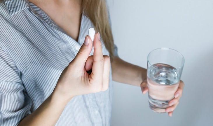 Na imagem, a mulher segura um comprimido em uma das mãos e um copo d'água na outra. Dúvidas com um ginecologista.