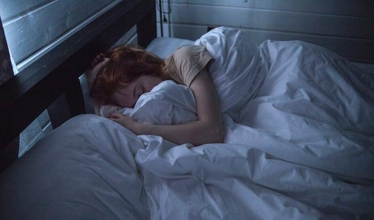 Na imagem, a mulher está deitada na cama com muitos cobertores como se sentisse dor. Dúvidas com um ginecologista.