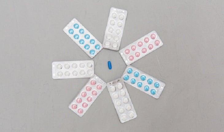 Na imagem, várias cartelas de remédio com comprimidos em rosa e azul estão dispostos em círculos com um comprimido azul no meio. Dúvidas com um ginecologista.