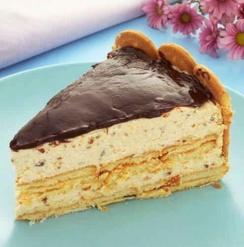 A foto contém a torta holandesa de biscoito maisena com amendoim, cobertura de chocolate, em um prato azul e flores flores ao fundo.