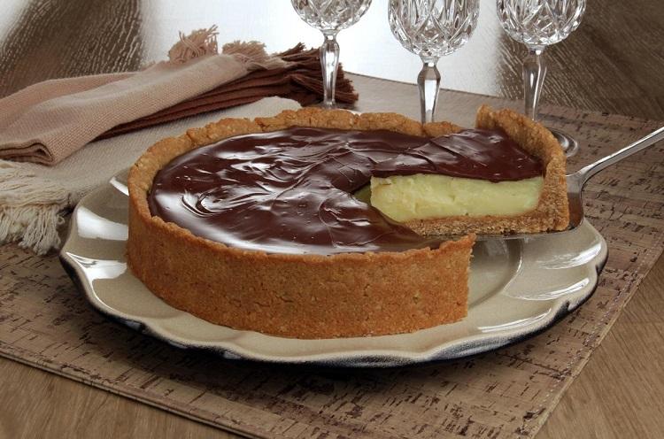 receita de torta de baunilha com ganache feita em uma fôrma de aro removível. A torta possui massa de biscoito, um creme branco e está coberta por um creme de chocolate.