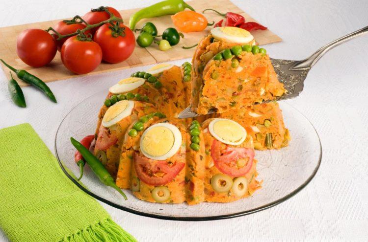 Cuscuz vegetariano, no prato transparente decorado com ovo cozido, ervilhas, azeitona, fundo com tomates e pimenta verde