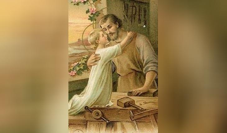 São José, com roupas de trabalho na cor bege, abraça o menino Jesus, que veste um manto branco e está sobre a mesa de trabalho de José