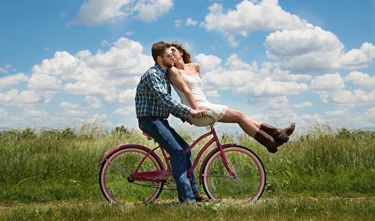 homem pedalando uma bicicleta e mulher sentada no guiidão dando um beijo em seu rosto