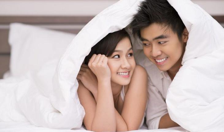 casal se olha sorrindo na cama, embaixo de um edredom branco