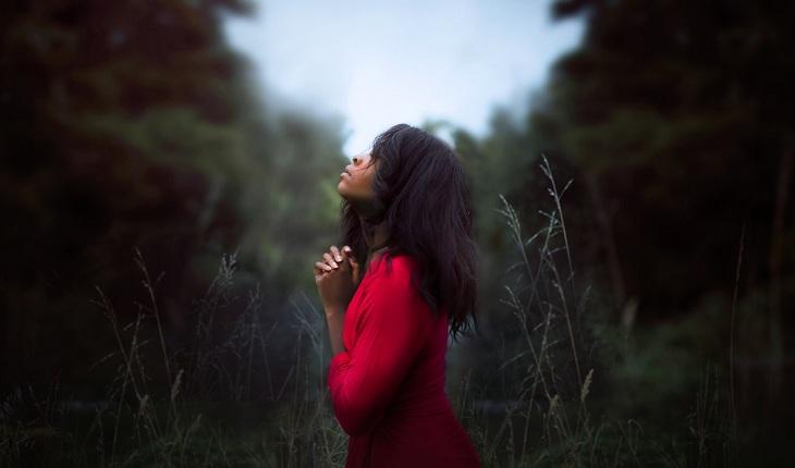 Na imagem, uma mulher está de vermelho no meio de árvores, na natureza, orando olhando para o céu. Novena de santa rita de cássia