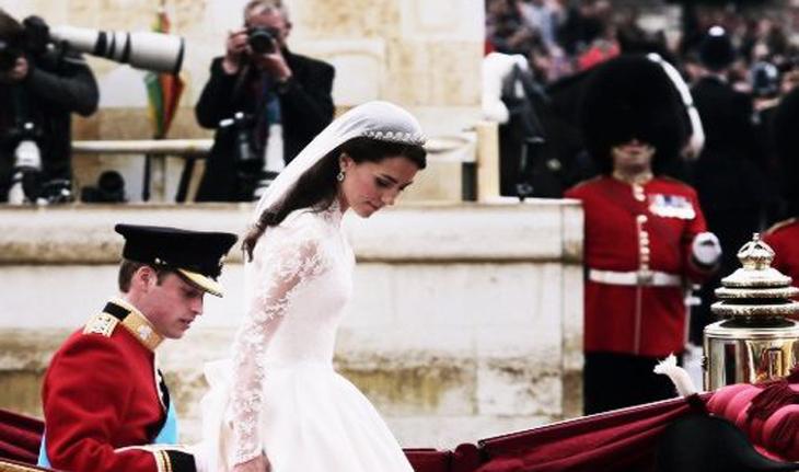 foto do casamento do príncipe William e Kate, eles esão saindo da carruagem na porta da igreja. Ela vente um vestido de noiva de manga comprida em renda e véu. Ele veste uma farda com muitas medalhas, quepe e luvas