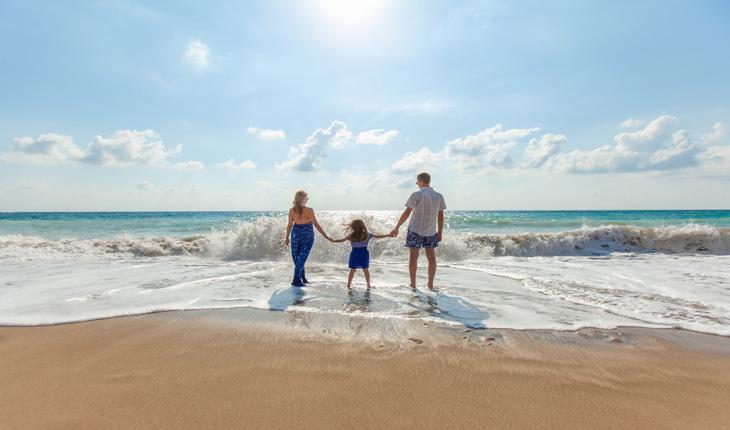 família com mãe, filha pequena e pai de mãos dadas em frente ao mar