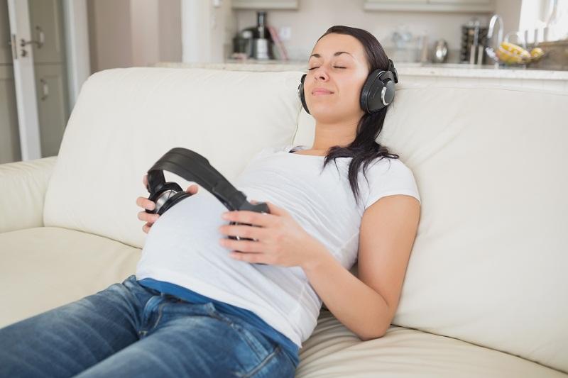 grávida de jeans e blusa branca, sentada no sofá com fone de ouvido na barriga, ouvindo música