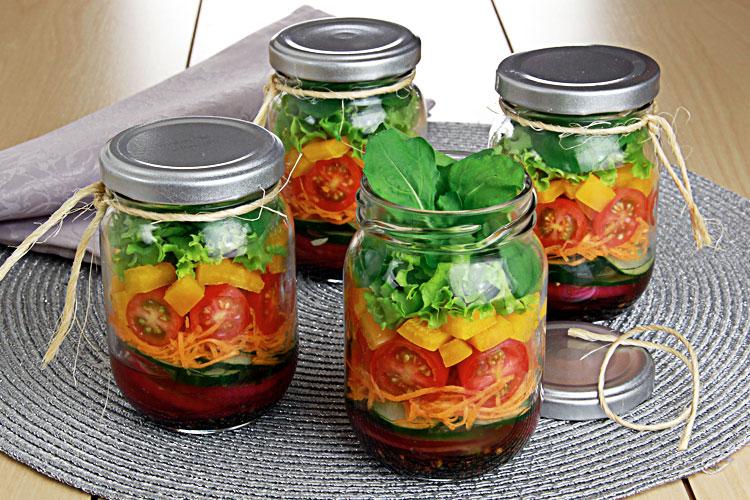 Quatro potes de vidro com saladas dentro. A receita conta com ingredientes como molho, tomate, cenoura e folhas verdes.