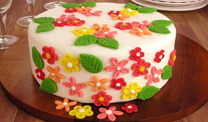 bolo decorado com pasta americana branca e flores, também feitas de pasta, nas cores rosa, salmão e amarelo com folhas verdes