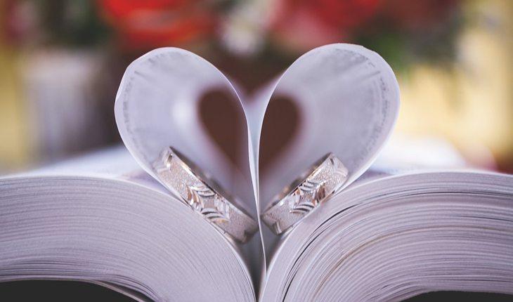 Na foto há um livro com as páginas formando um coração e dentro dele há dua alianças de compromisso.