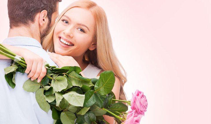 Na foto há uma casal abraçado. O homem está de costas e a mulher de frente, olhando para a câmera, e segurando um ramalhete de flores.