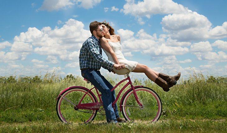 Na foto há um homem andando de bicicleta e uma mulher na parte do guidão, sentada. Ela está virada para trás beijando a bochecha do homem.