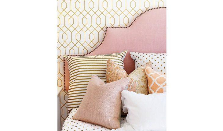 Na foto é possível ver o canto de uma cama com uma cabeceira rosa e almofadas e travesseiros com estampas da mesma tonalidade ou em cores próximas