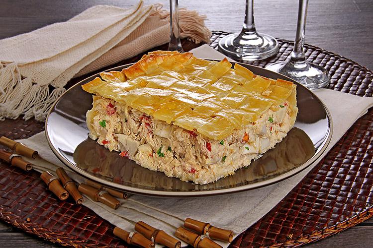 Pedaço quadrado de torta em um prato de servir com guardanapo e taças ao fundo.