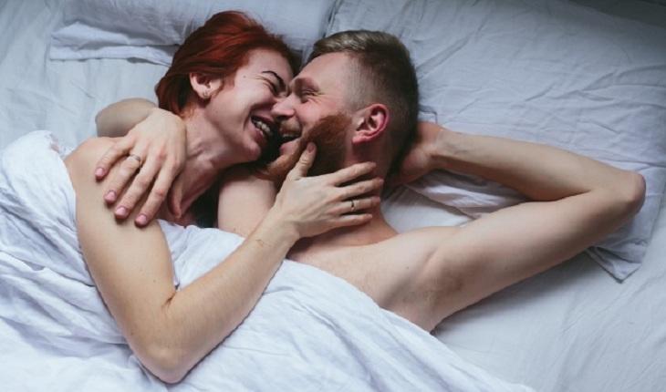Na imagem, o casal está deitados juntos, rindo e se rocando, nus na cama com cobertor. Prática carinhosa e intensa.