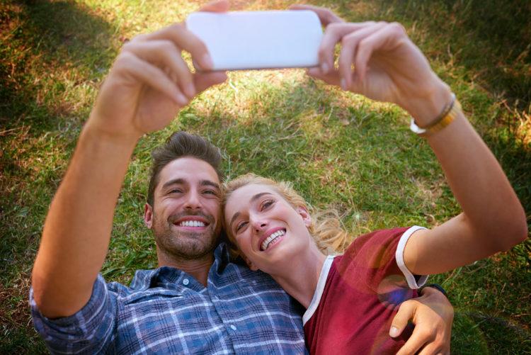 Casal, deitados no gramado, sorrindo, tirando selfie com celular, aplicativos