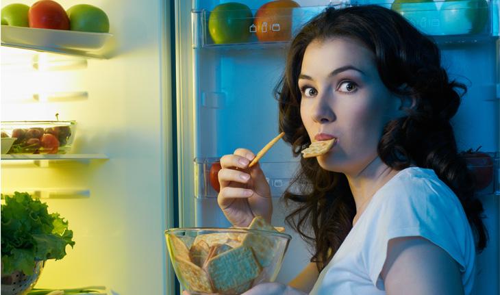 Mulher comendo bolacha à notie de frente para uma geladeira com comidas saudáveis