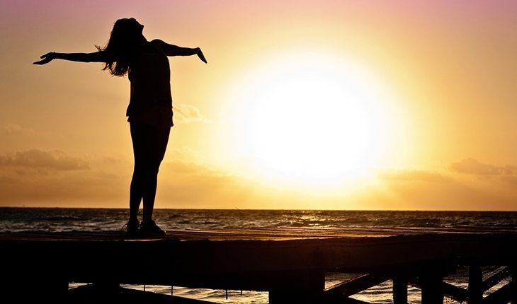 Na foto há uma mulher de braços aberto na beira de uma praia no pôr do sol