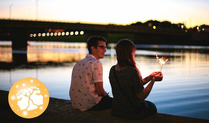 Na foto há um casal heterossexual sentado em uma ponte no pôr do sol.